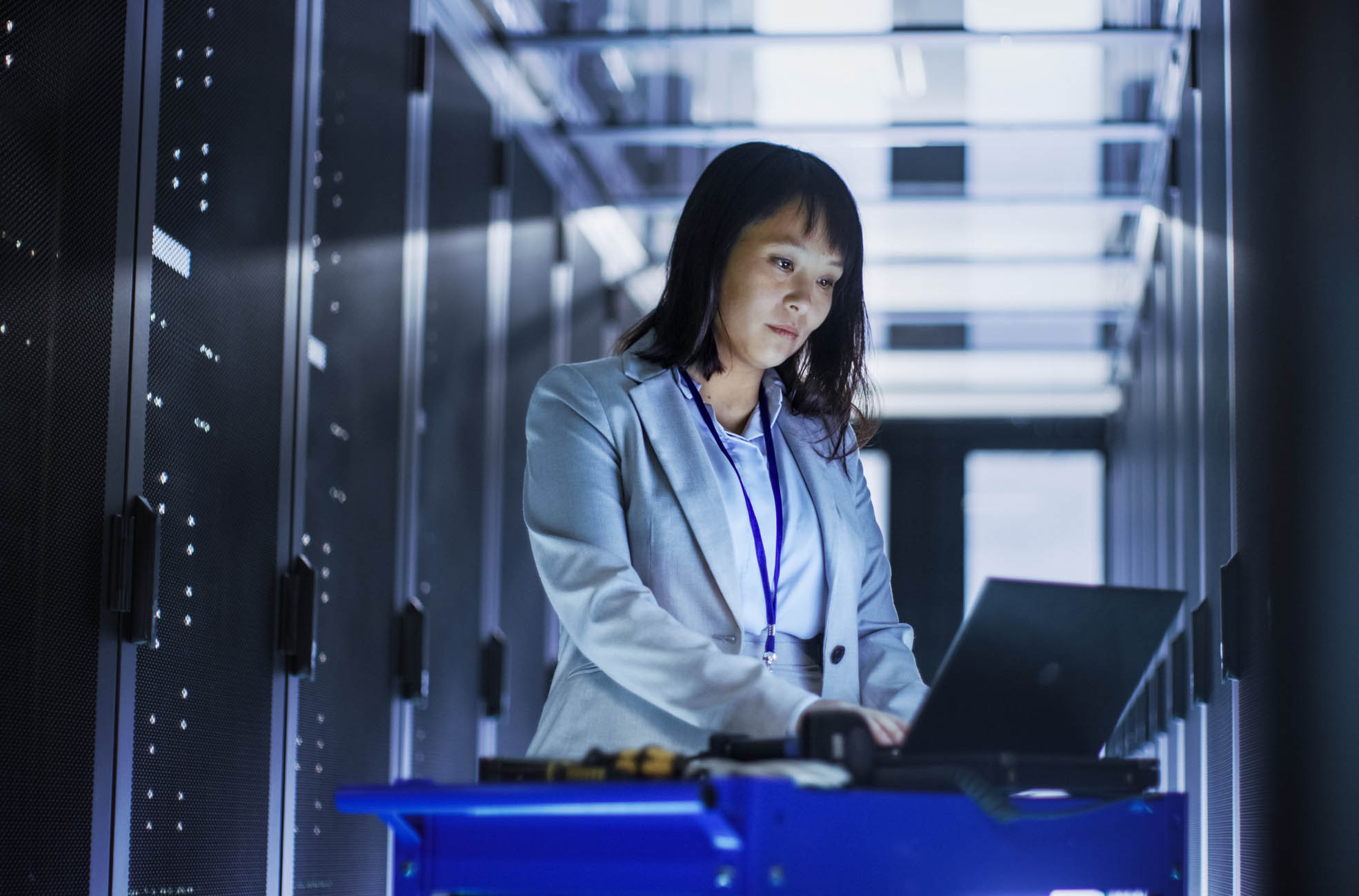 Asian Female IT Engineer Working on a Laptop on Tool Cart, She Scans Hard Drives.  She’s in a Big Data Center Full of Rack Servers.